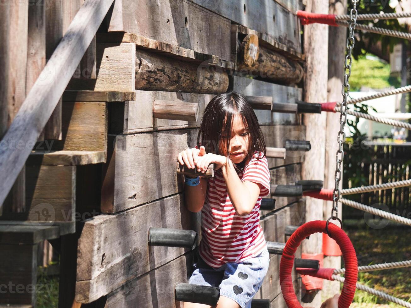 Children play outdoors on a wooden playground in a park. Happy girl playing on wooden climbing wall in playground during summer. photo
