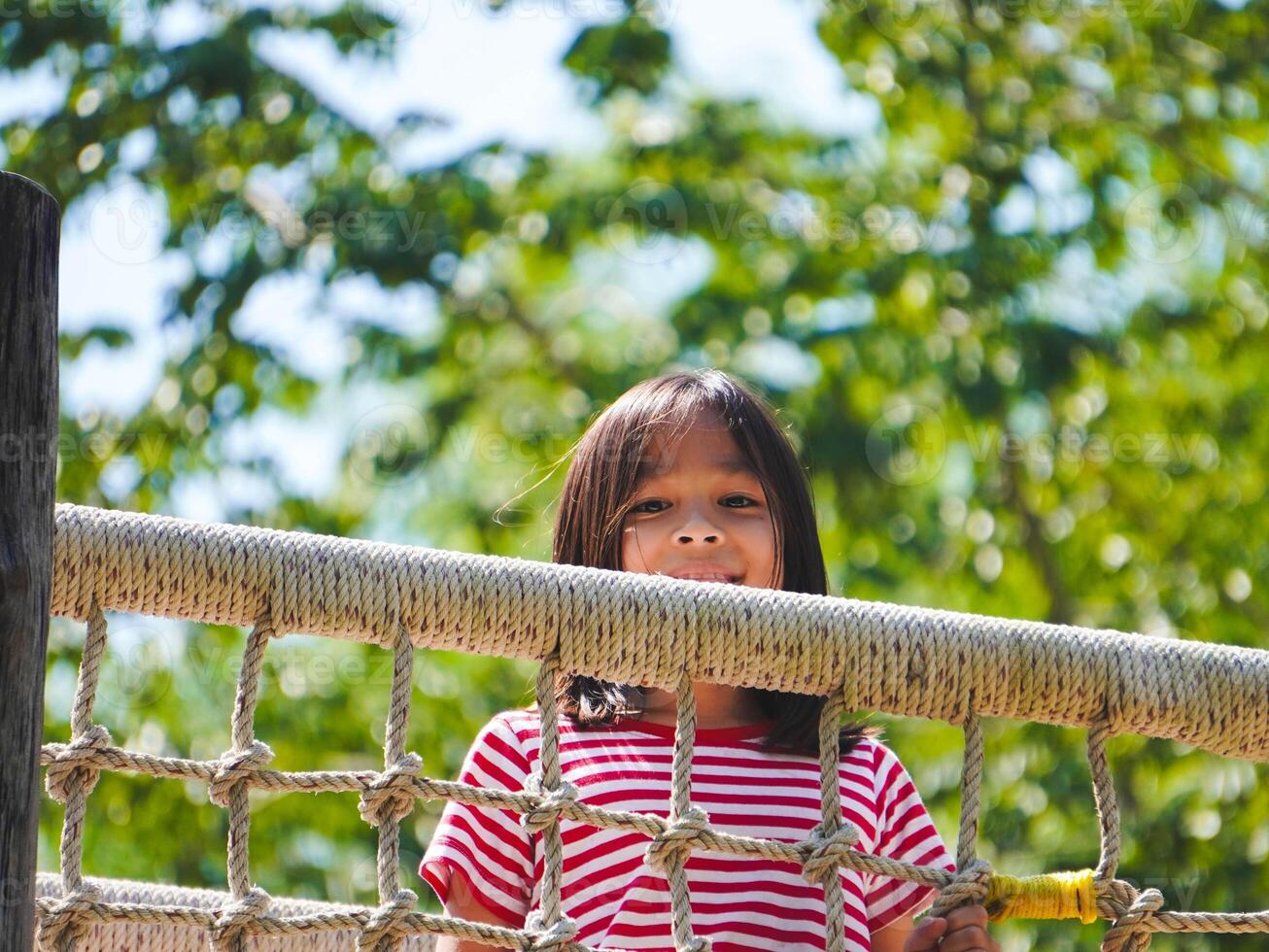Children play outdoors on a wooden playground in a park. Happy girl playing on a rope net bridge in playground during summer. photo