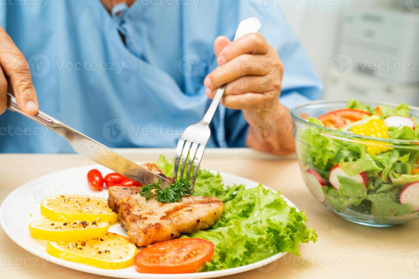 Asian senior woman patient eating pork chop stake and vegetable salad for healthy food in hospital. photo