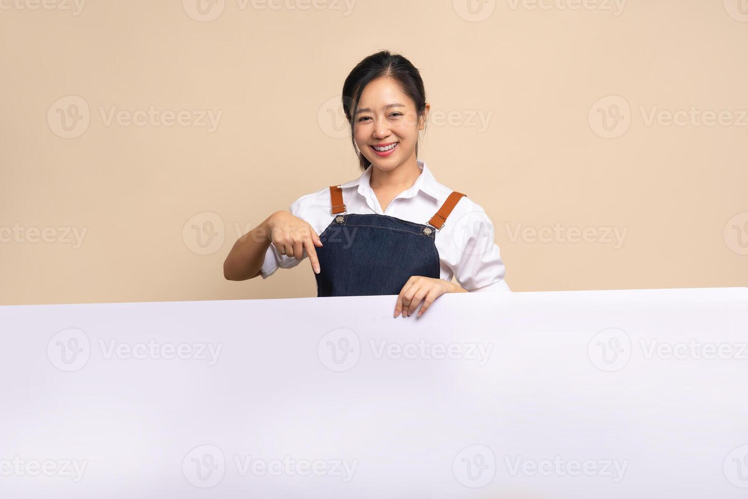 Cute Asian woman wearing apron with a smile. She is pointing her finger at your ad space on a beige background. photo