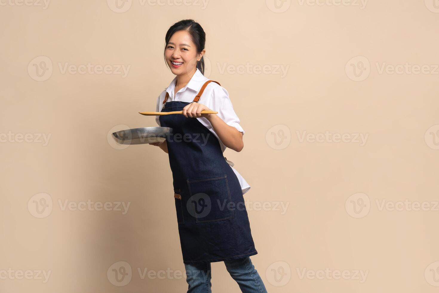 Beautiful and attractive Asian female coffee shop owner wearing apron. She holds a frying pan and a wooden spatula on a beige background. photo