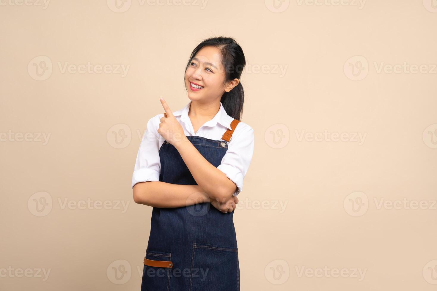 Beautiful smiling Asian female entrepreneur in apron standing isolated on beige background. She points her finger at the space for your advertisement. photo