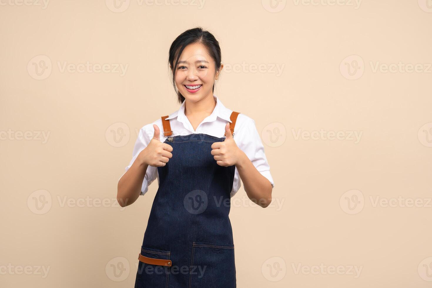 Portrait smiling young asian woman wearing apron showing thumbs up with hand isolated beige background photo