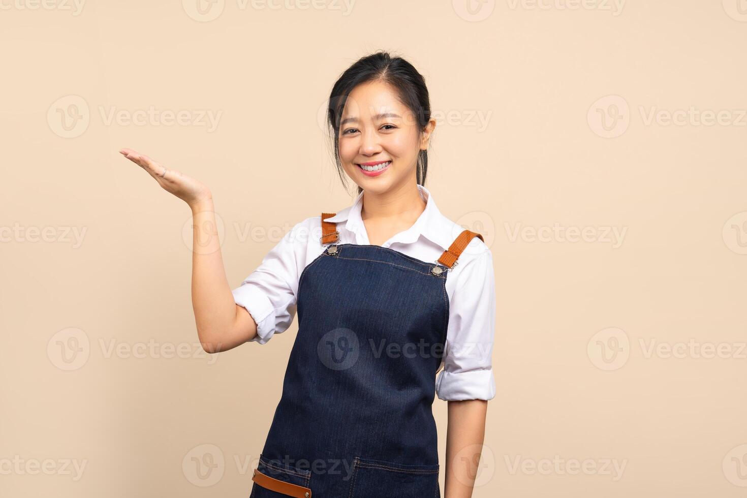Beautiful Asian woman in apron pointing at the empty space for your ad on beige background photo