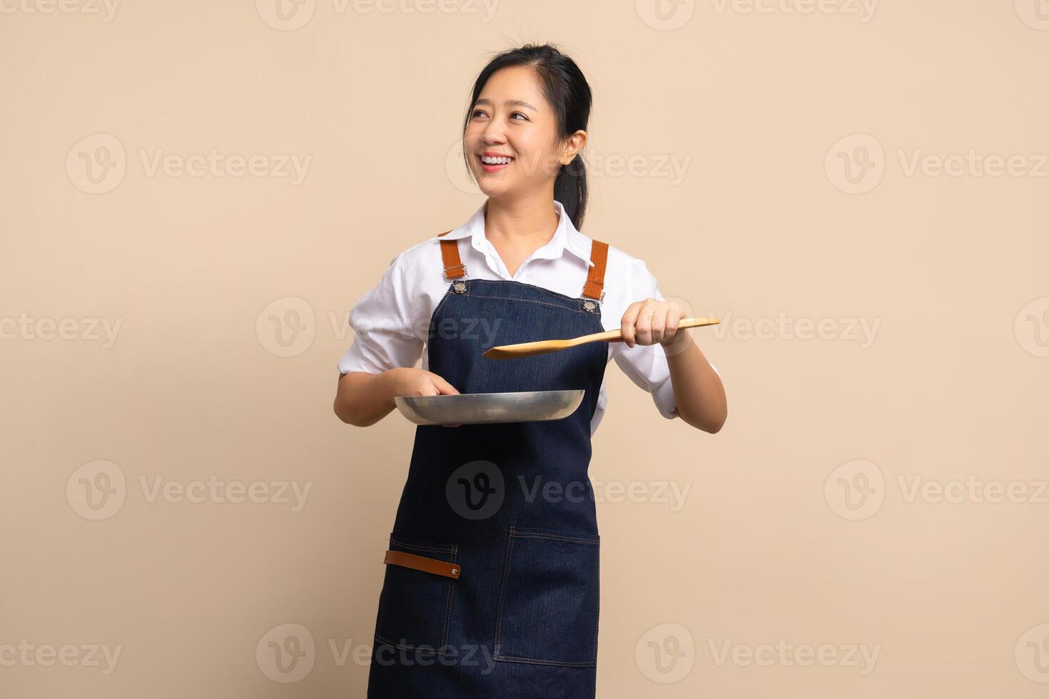 Beautiful Asian female coffee shop owner wearing apron. She holds a frying pan and wooden spatula with copy space. photo