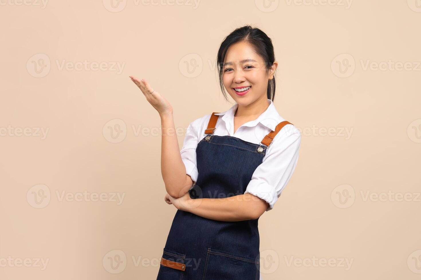 Cute Asian woman in apron with a smile pointing her finger to show space for your ad on beige background. photo