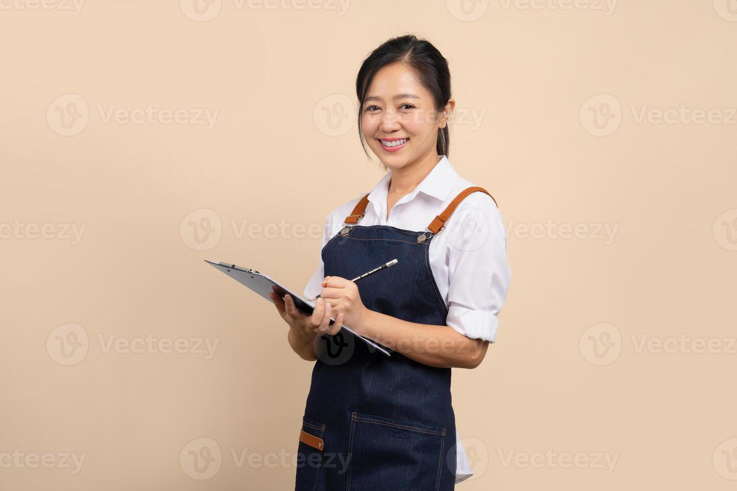 Smiling Asian female business owner wearing apron and holding tablet taking orders on beige background photo