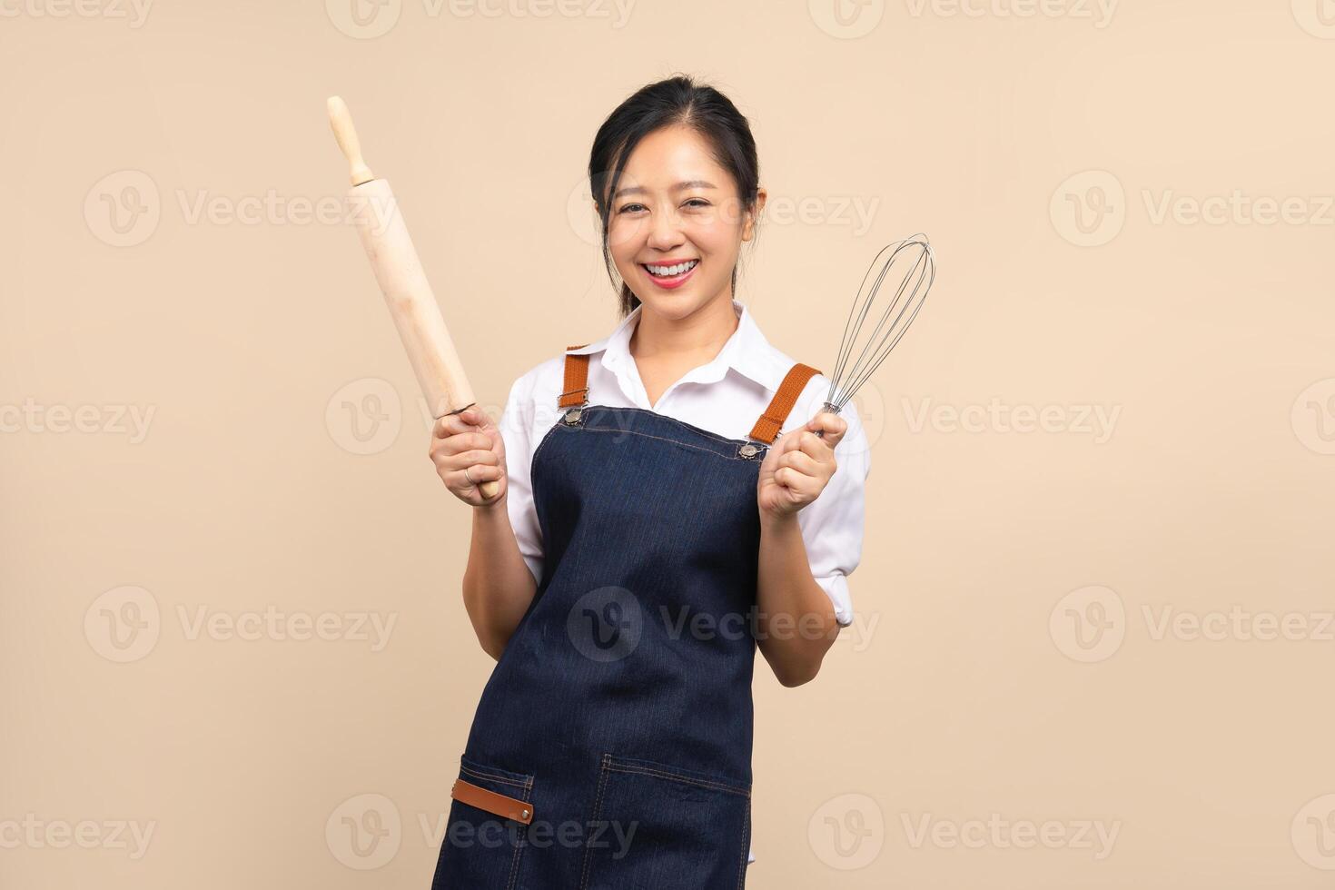 Beautiful Asian woman in apron holding kitchen utensils isolated on beige background photo