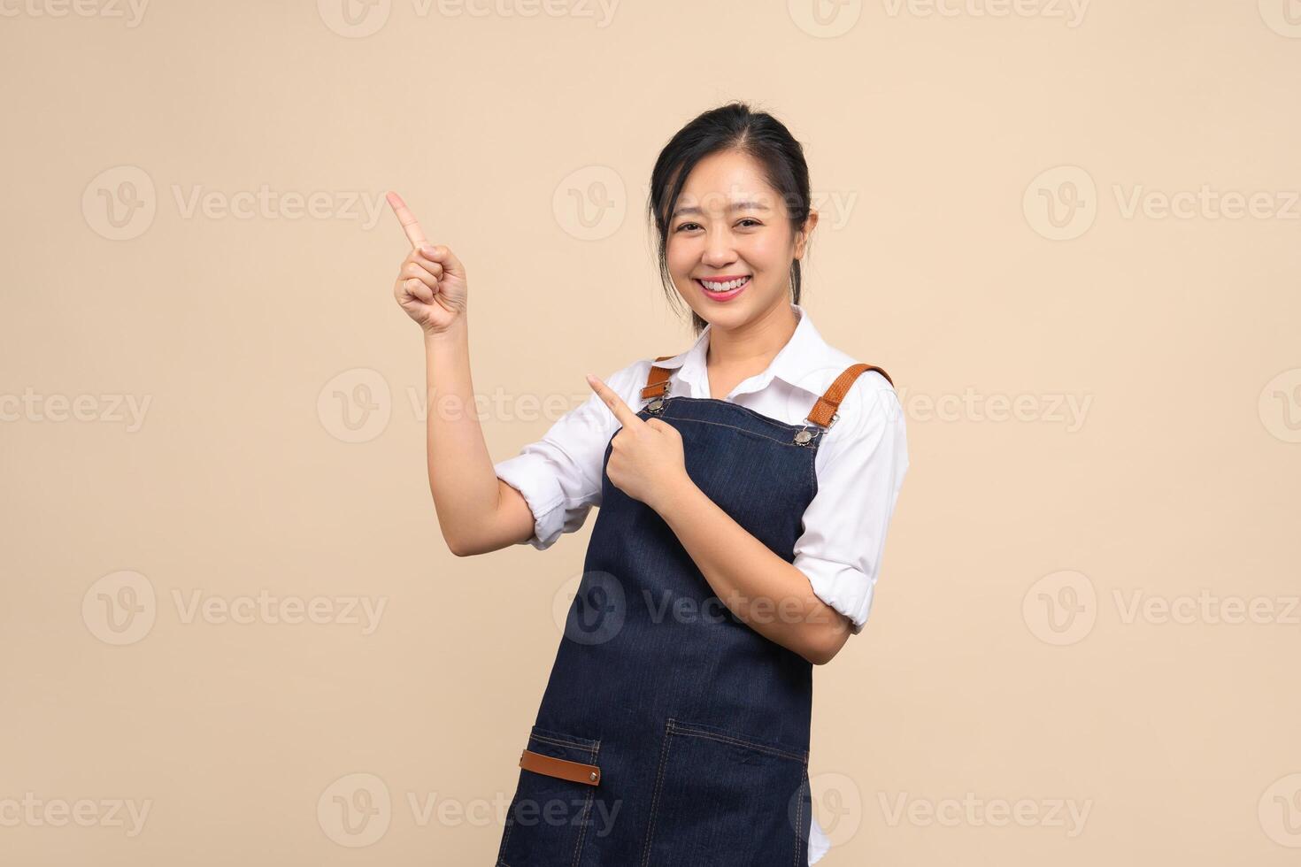 Cute Asian woman in apron pointing her finger to show space for your ad on beige background. photo