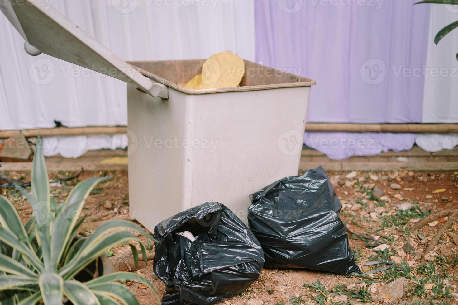 A garbage bin with two black trash bags placed beside it photo