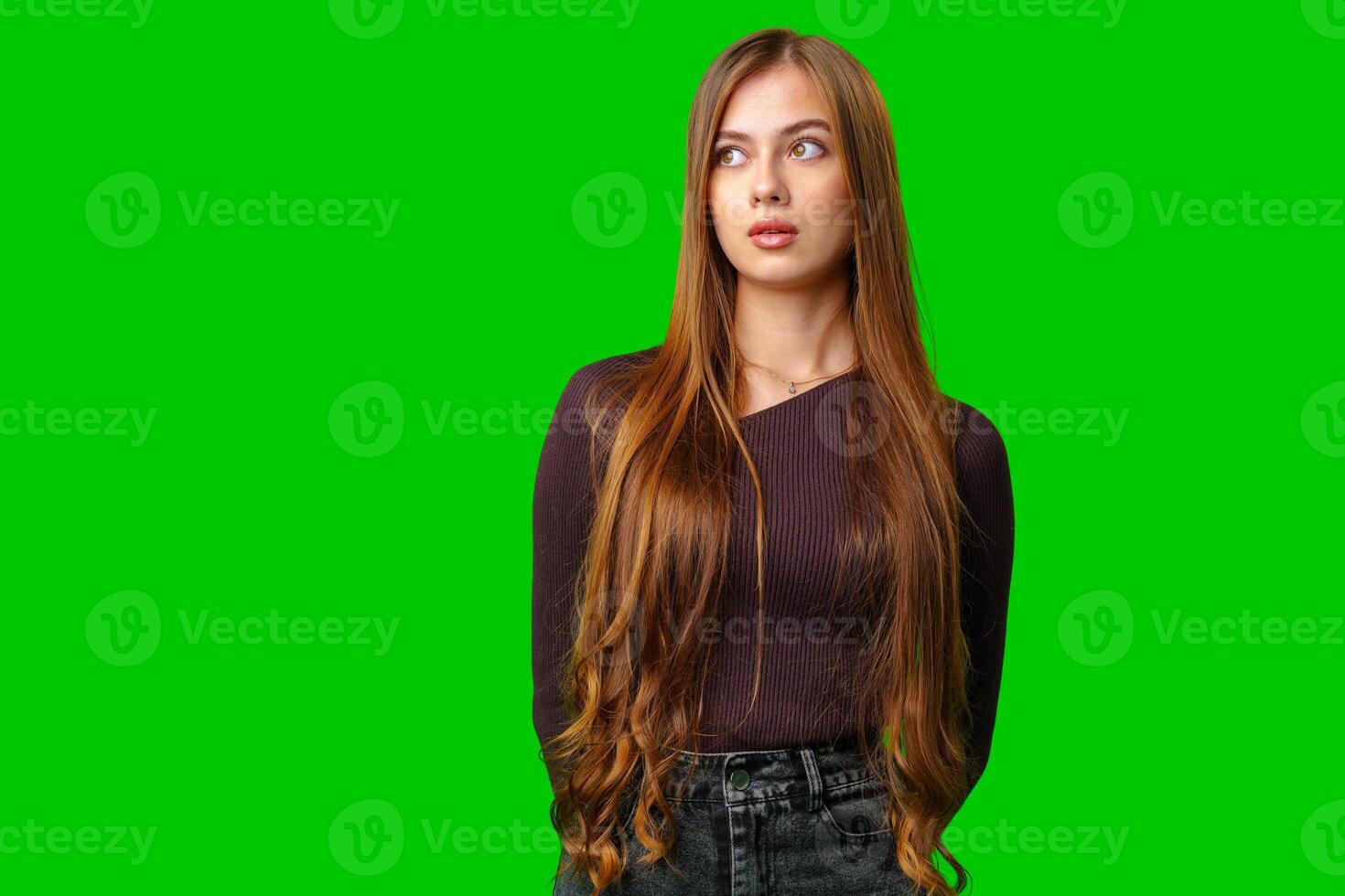 Young Woman With Long Brown Hair Looks Up and to the Right While Wearing a Purple Sweater and Jeans Against a Green Screen photo