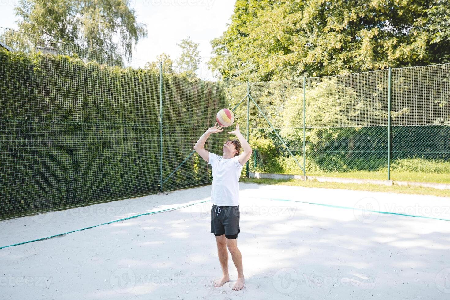 Young man tossing a volleyball into the air on a sandy court during a sunny day. The casual outdoor scene highlights fun, summer sports, and recreation in a natural setting photo