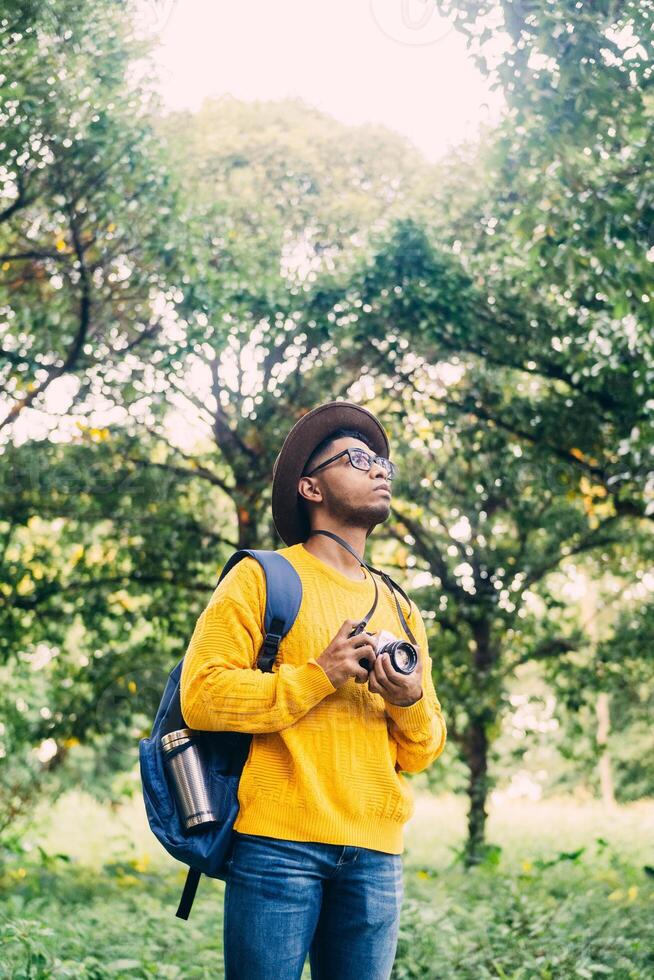 Young Latin American man in a green forest is wearing a hat, glasses and a yellow sweater holding the camera. photography and travel concept. photo
