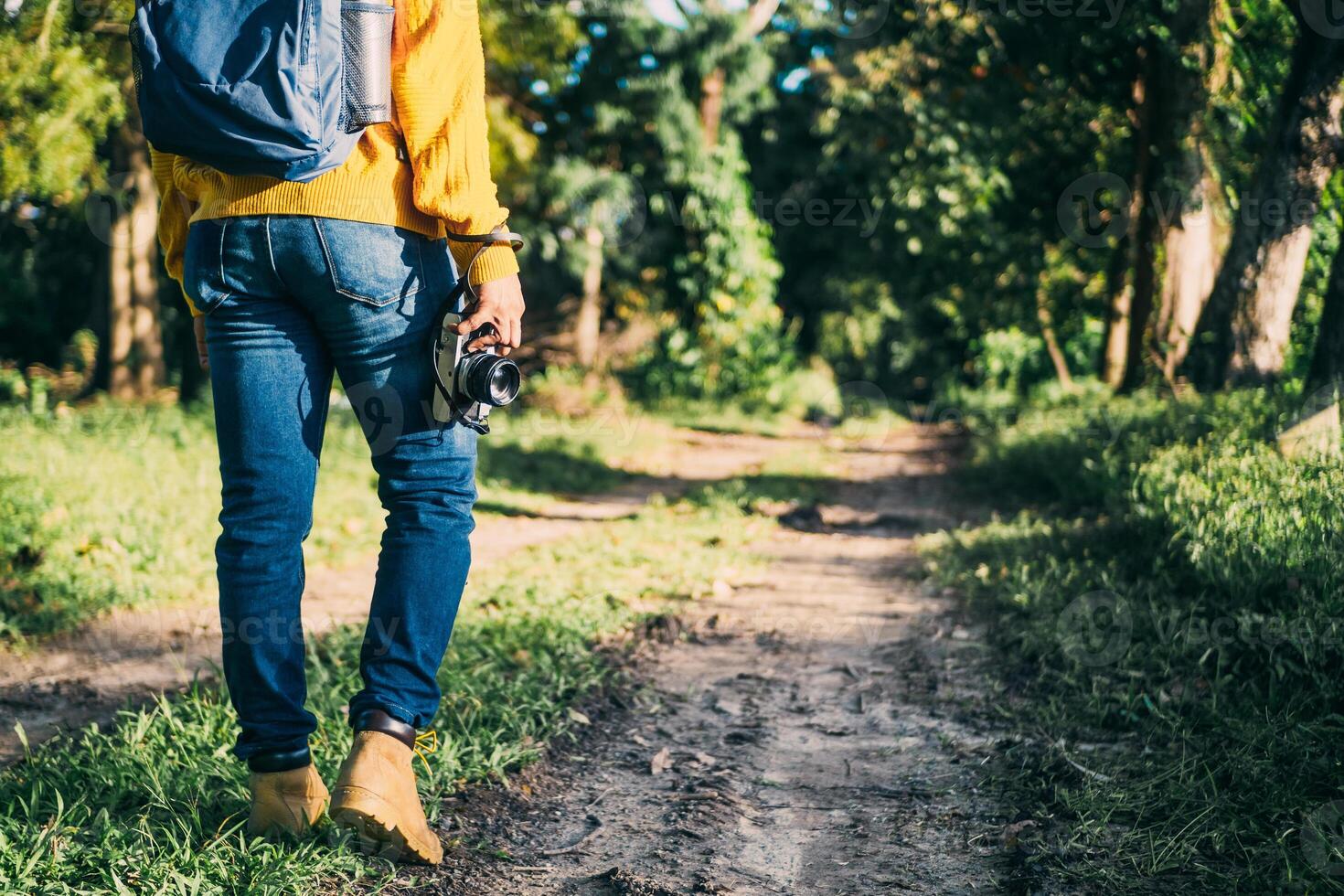 unrecognizable man with backpack and boots holding a camera in front of a path in a forest, nature travel and hiking concept. photo