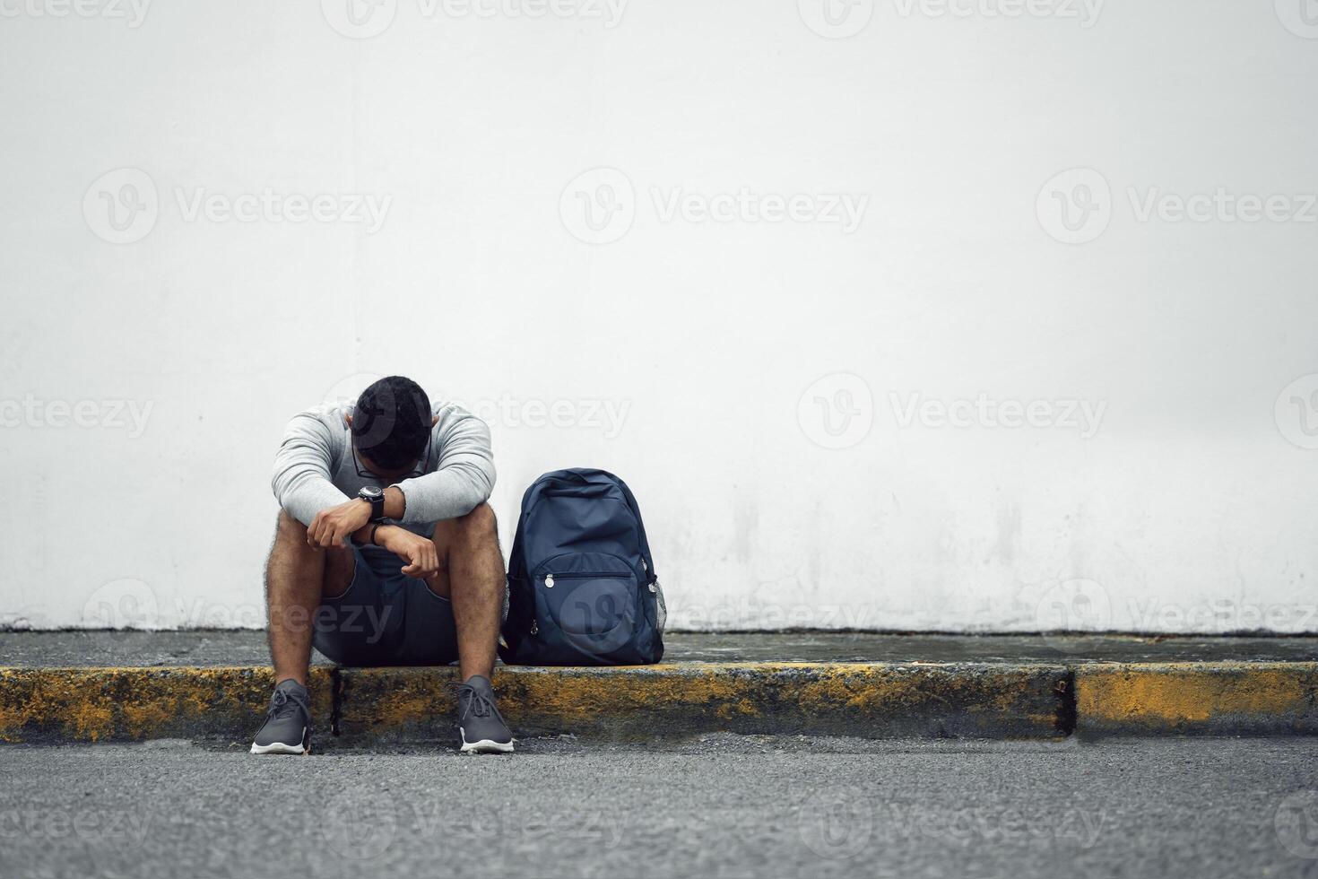 young man hispanic sitting on the sidewalk of a street in front white wall with a lost look of disappointment and sadness, depression concept. photo