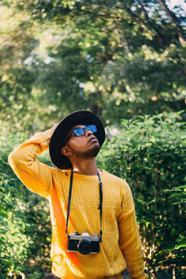 Young Latin American man in a forest wears a yellow sweater with a camera and a hat, looking around. travel and natural recreation concept. photo