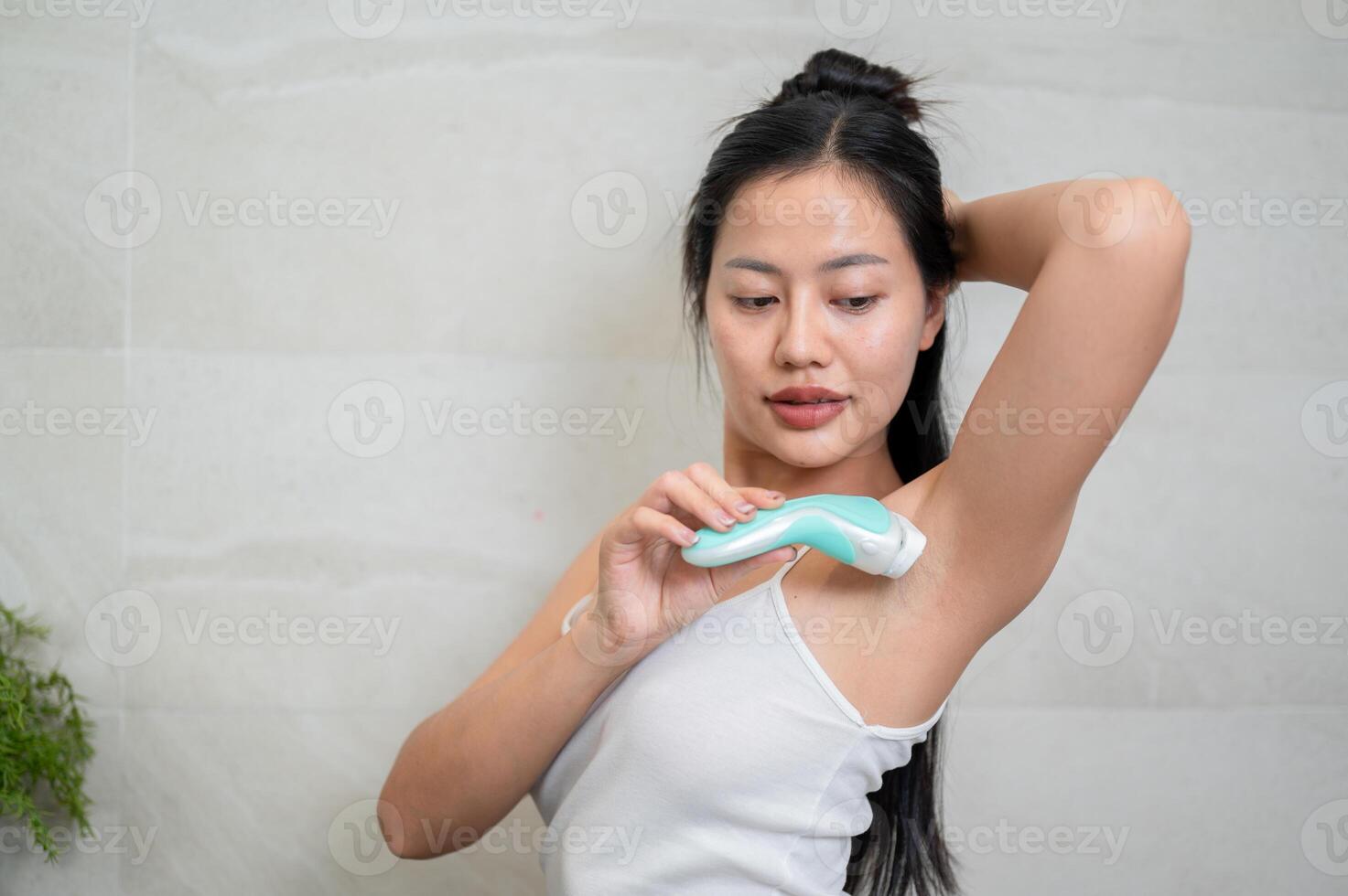 A beautiful Asian woman is shaving her armpit with a feminine razor in front of the mirror. photo