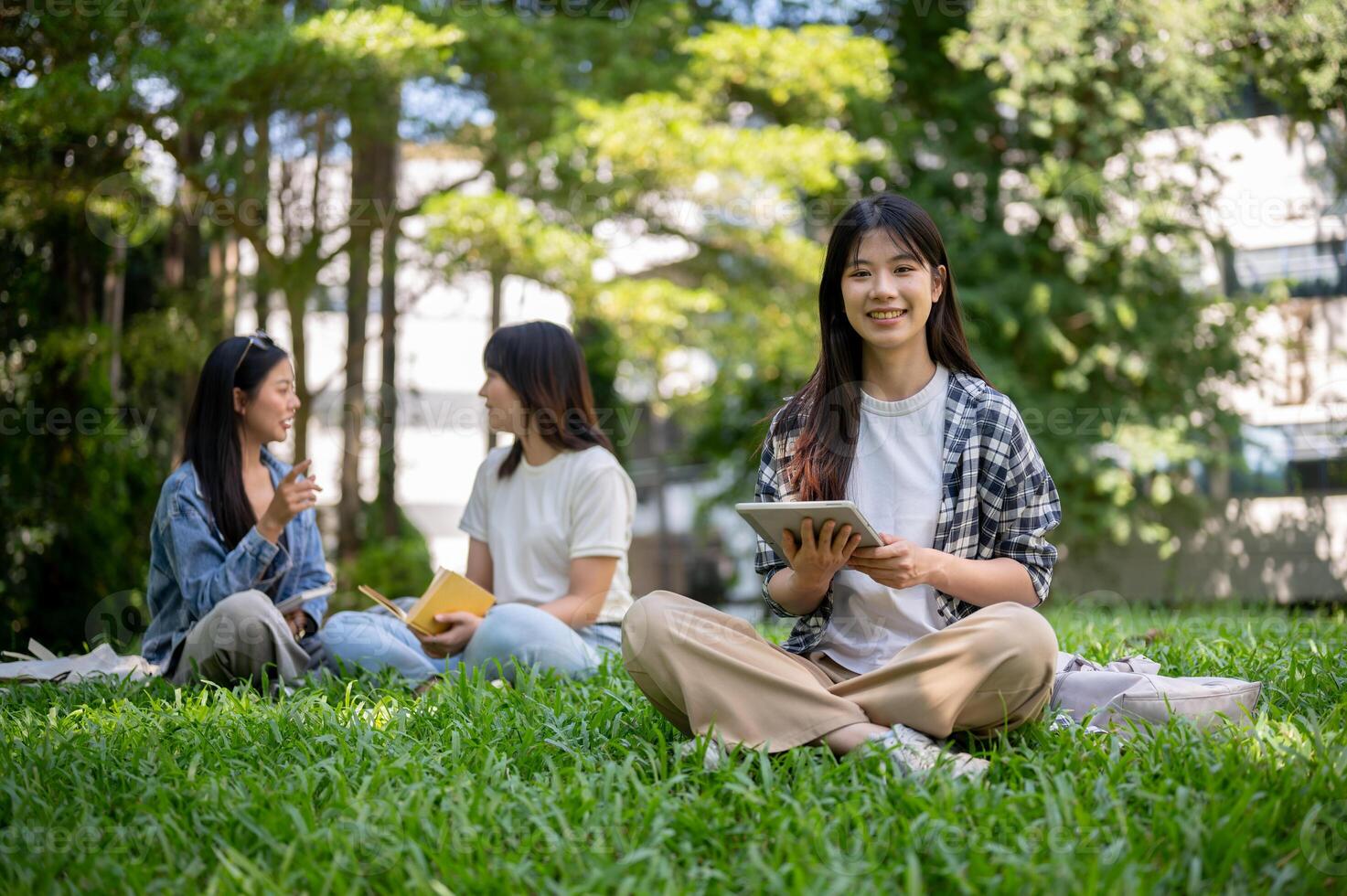 Charming Asian female college student sits on the grass in her college campus park, holding a tablet photo