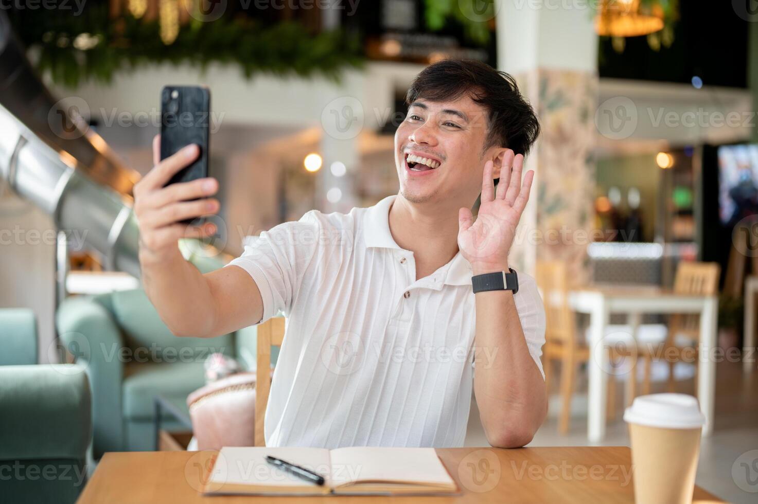 A cheerful Asian man is enjoying a call with his friend on his smartphone in a coffee shop. photo