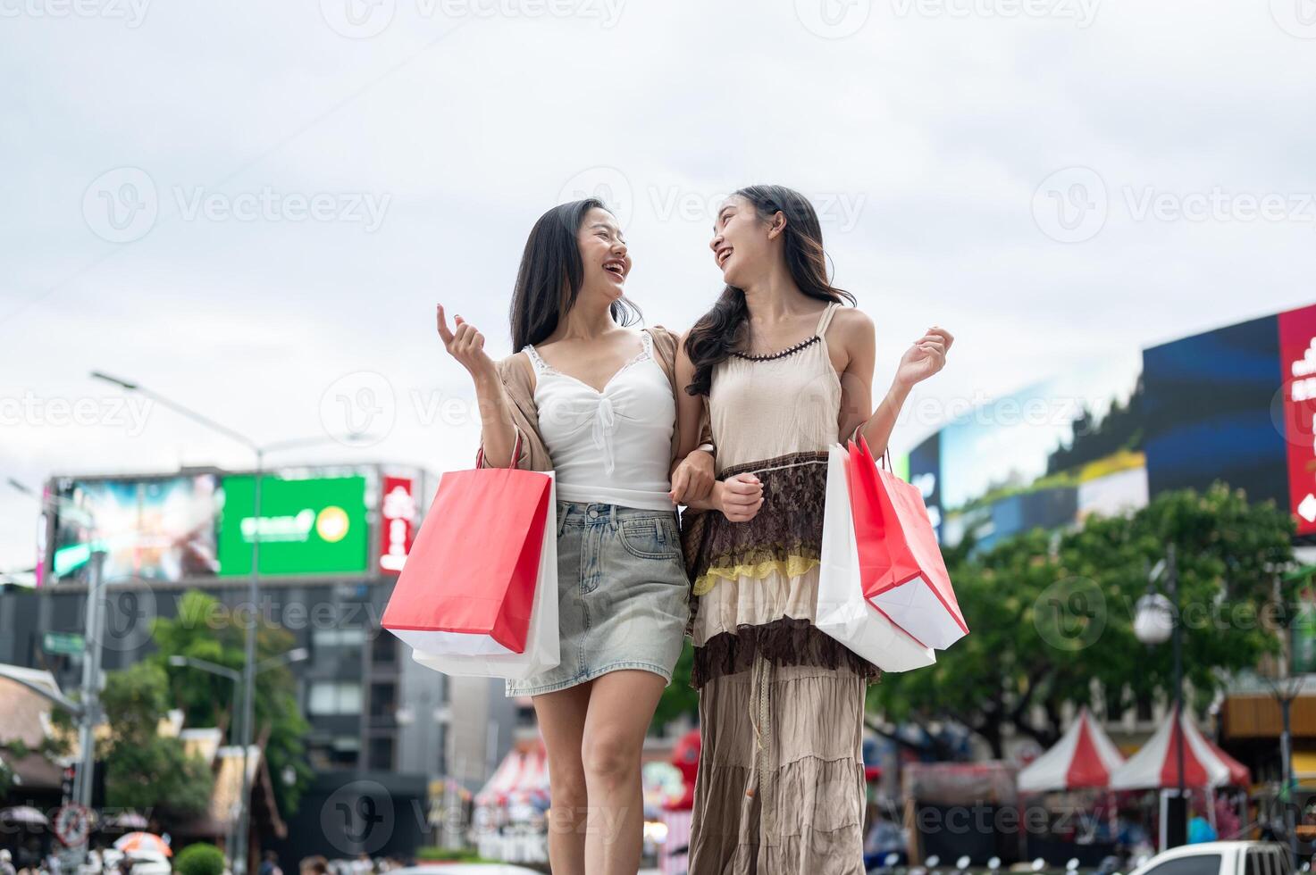 Two attractive Asian female friends smile at each other, carrying their shopping bags by a street. photo