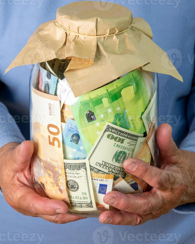 Close-up image of a persons hands holding a glass jar filled with various Euro and US dollar bills photo