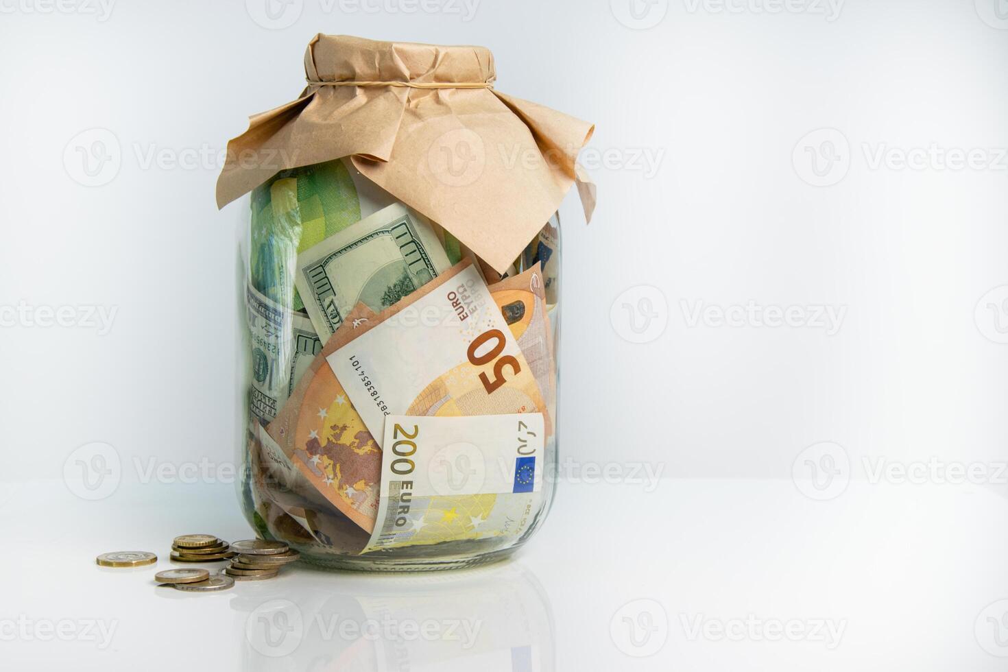A glass jar filled with US dollar and Euro bills with a few coins scattered around it photo