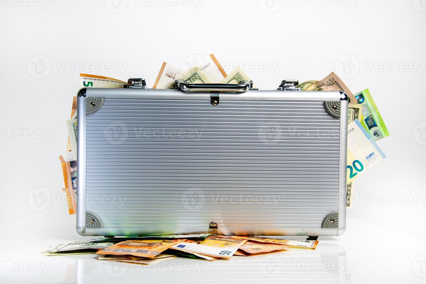 A silver briefcase sits on a white surface with Euro and US Dollar bills spilling out of it photo