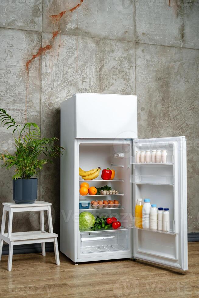 White Refrigerator With Open Door Showing Food Inside, Grey Concrete Wall, Wooden Floor, and Plant photo