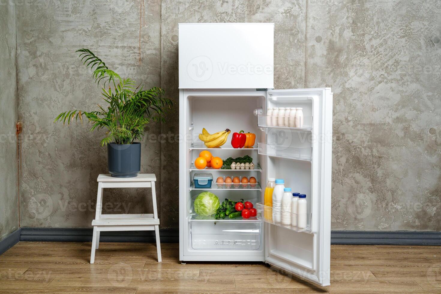 White Refrigerator With Open Door Showing Food Inside, Grey Concrete Wall, Wooden Floor, and Plant photo