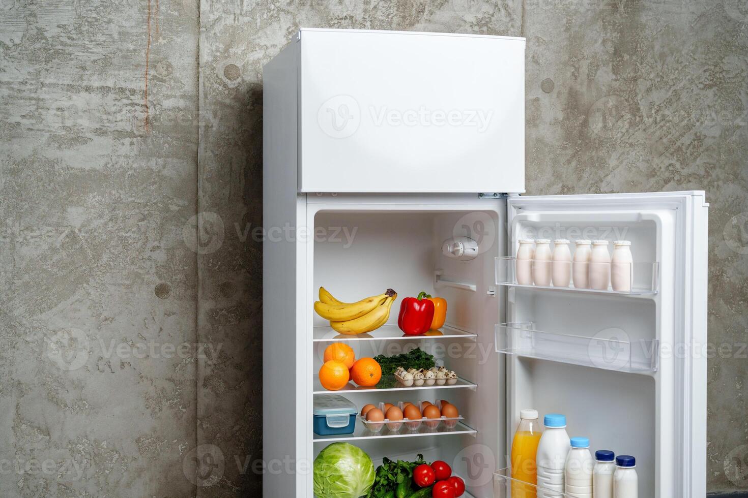White Refrigerator With Open Door Showing Food Inside, Grey Concrete Wall, Wooden Floor, and Plant photo
