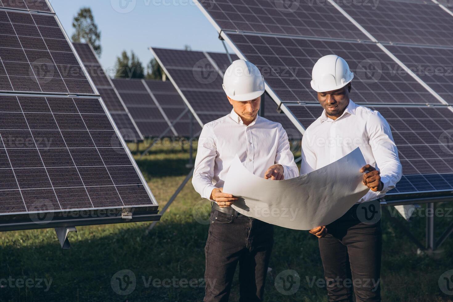 Two Engineers Wearing Hard Hats Analyzing Blueprints at a Solar Panel Field on a Sunny Day photo