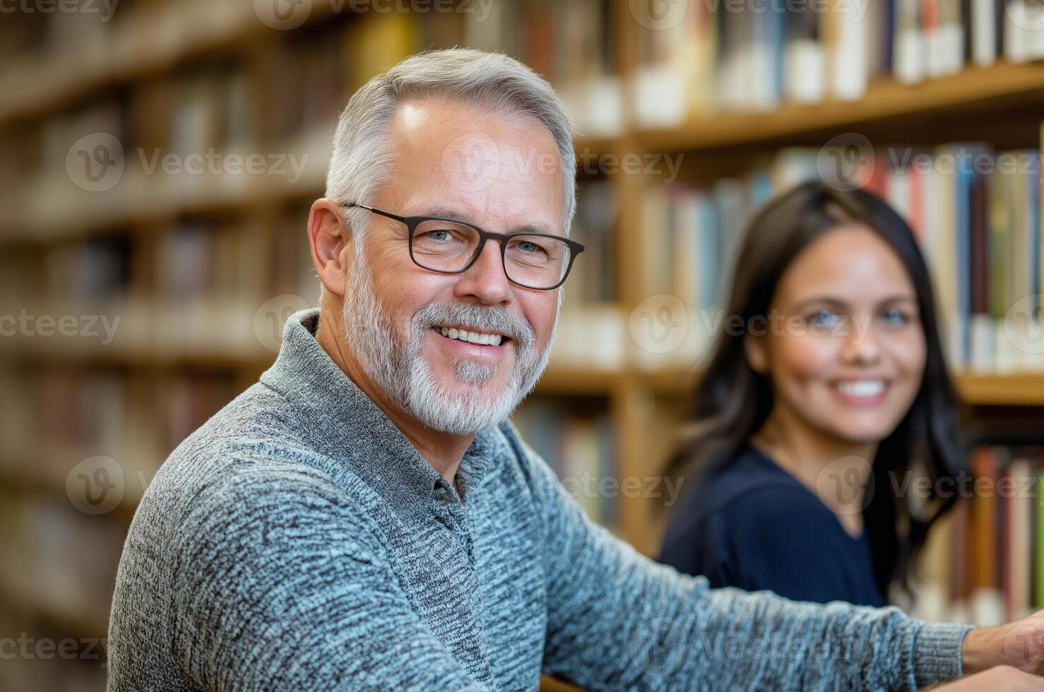 Middle-aged man smiling at the camera while reading in a library with a young woman in the background photo