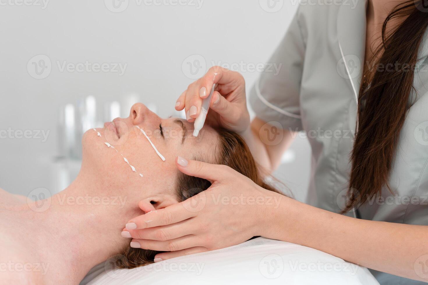 Masseuse applying cream with tube on woman's face at cosmetology salon photo