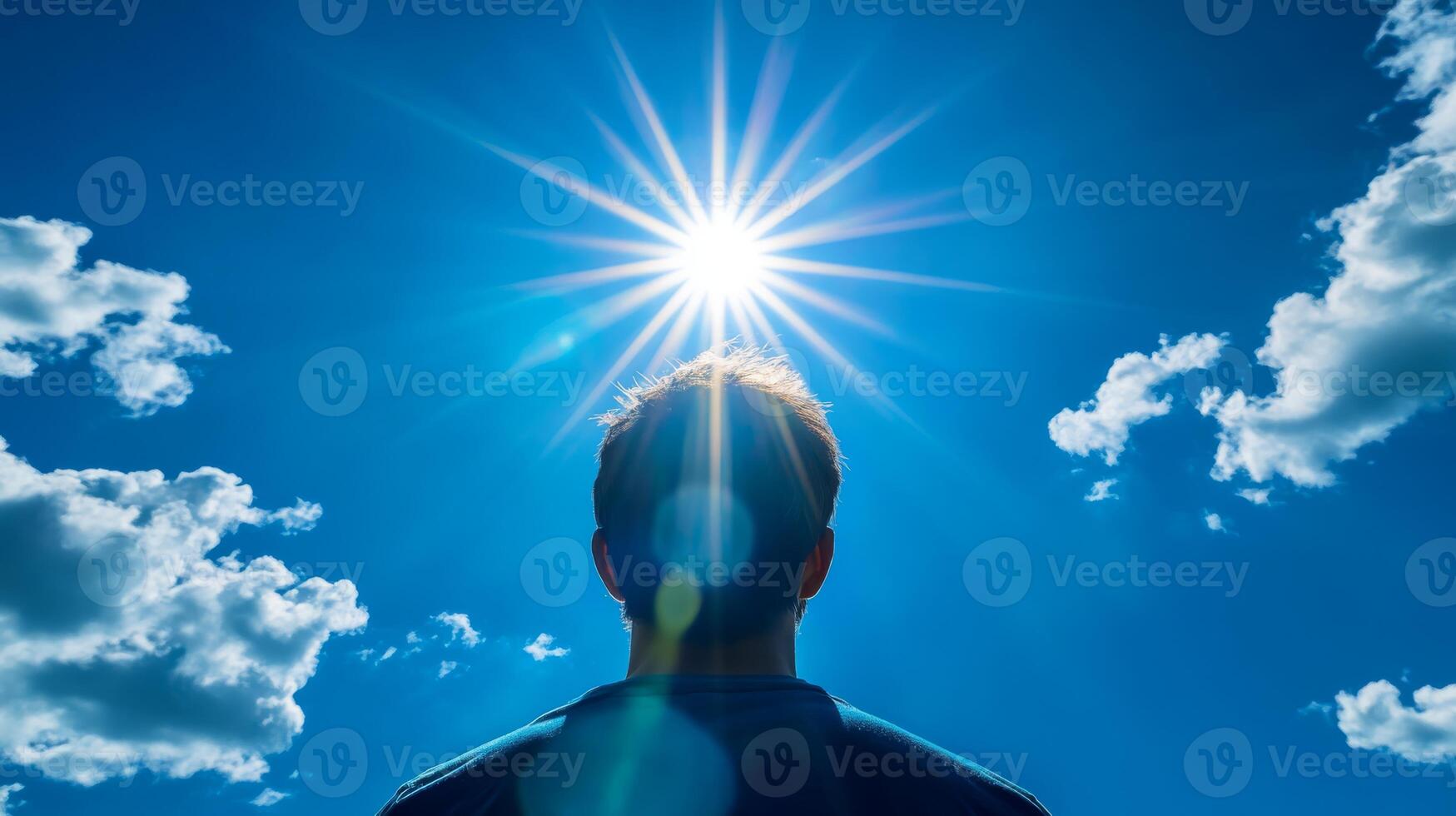 A striking silhouette of an individual uniquely stands against the backdrop of a bright sun, with its golden rays spilling over a clear blue sky, while fluffy white clouds drift gently by photo
