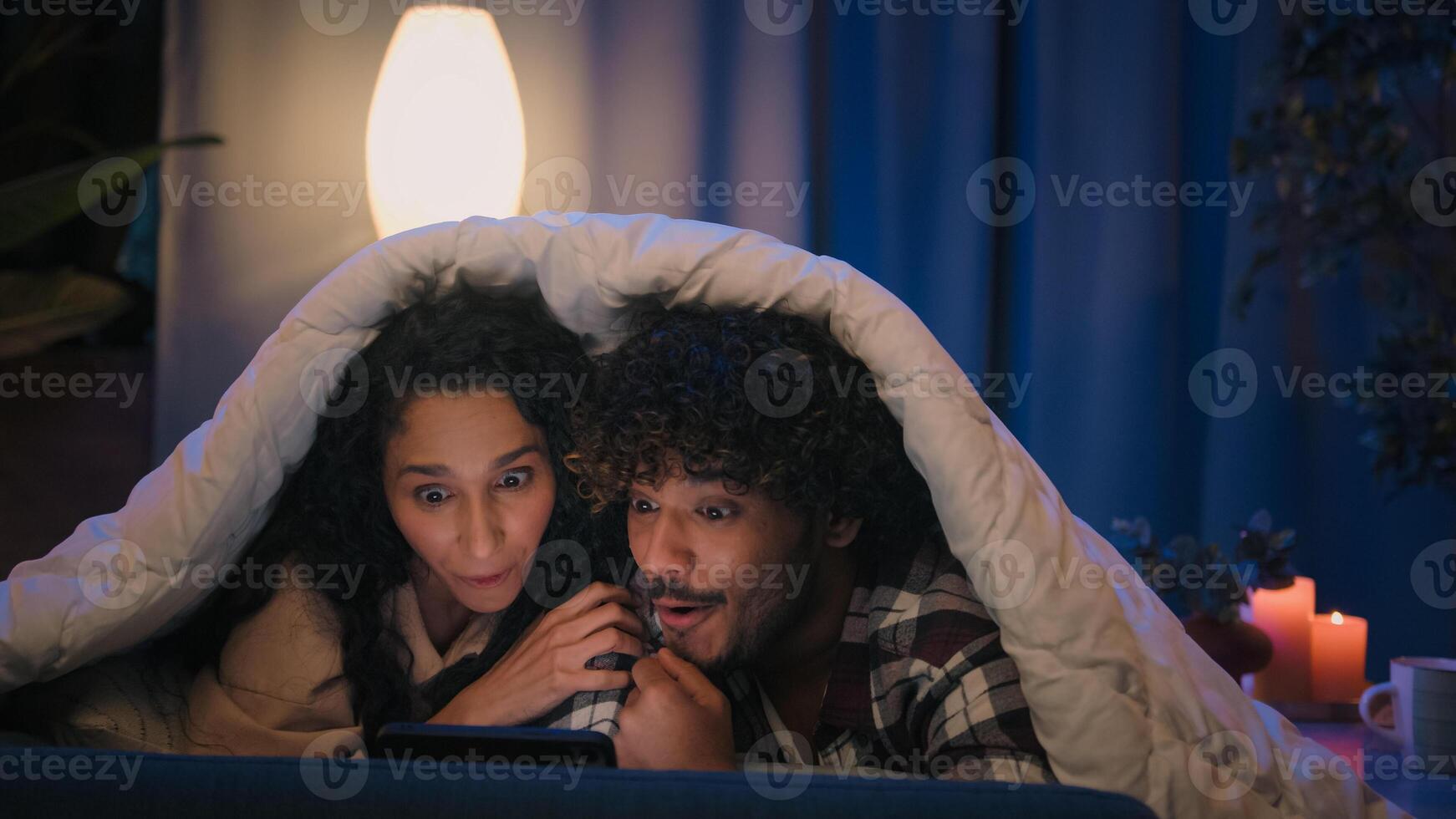 Happy family relaxed multiracial couple Indian man and Arabian woman at evening late night in home bed under blanket watching movie film with mobile phone talking discussing emotional amazed photo