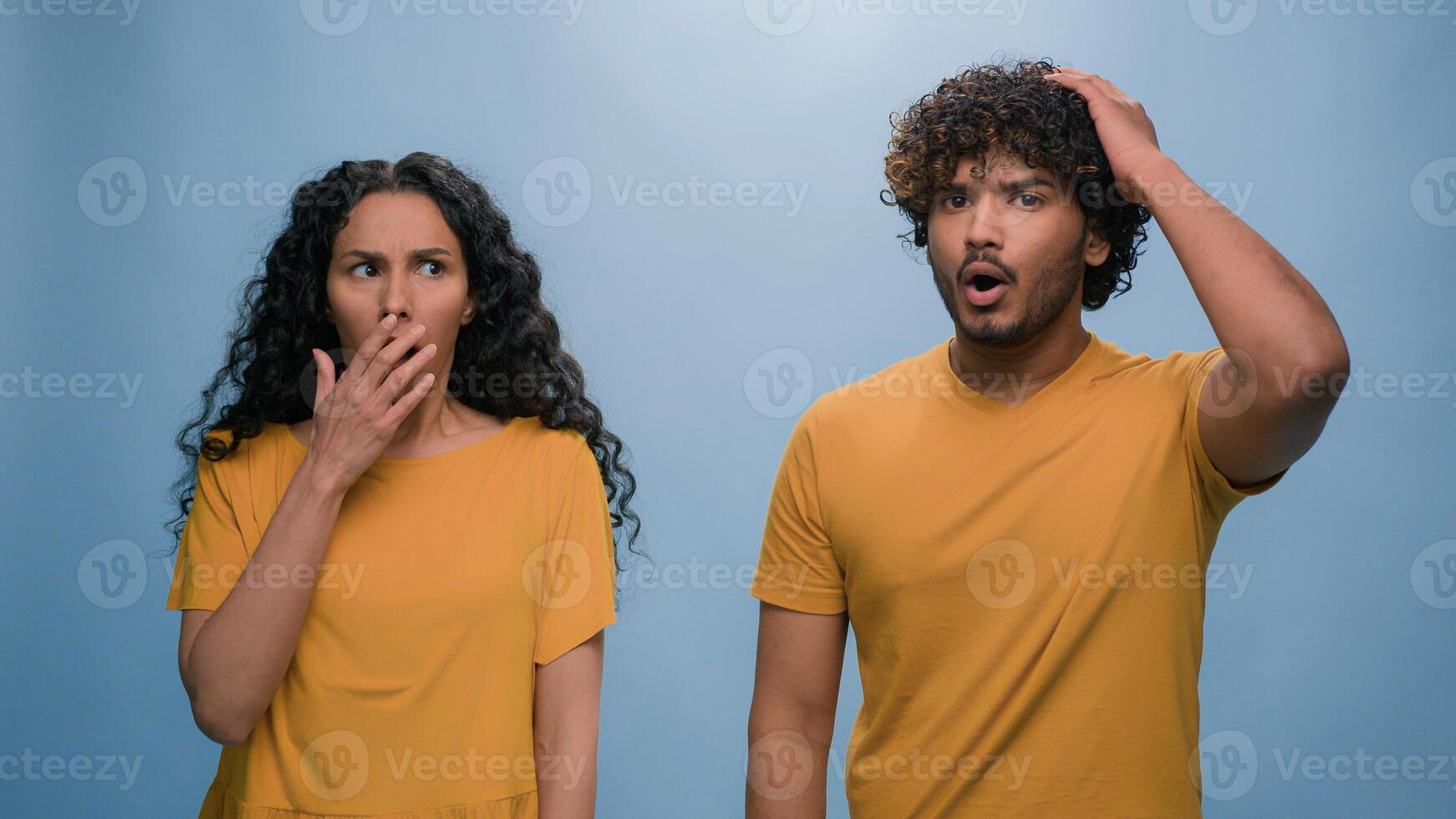 Multiracial shocked scared afraid couple amazed shocked Hispanic woman hiding behind Indian man astonishment wonder scare terrible horror ugly reaction blue studio background shock astonish fright photo