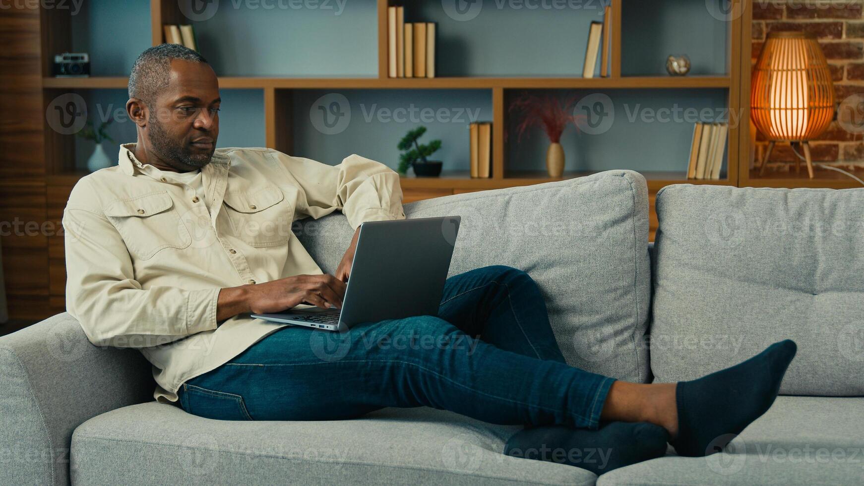 Mature african american man lying on sofa relax using computer businessman freelancer typing on laptop work internet at home ethnic male entrepreneur browsing social network send email online chatting photo