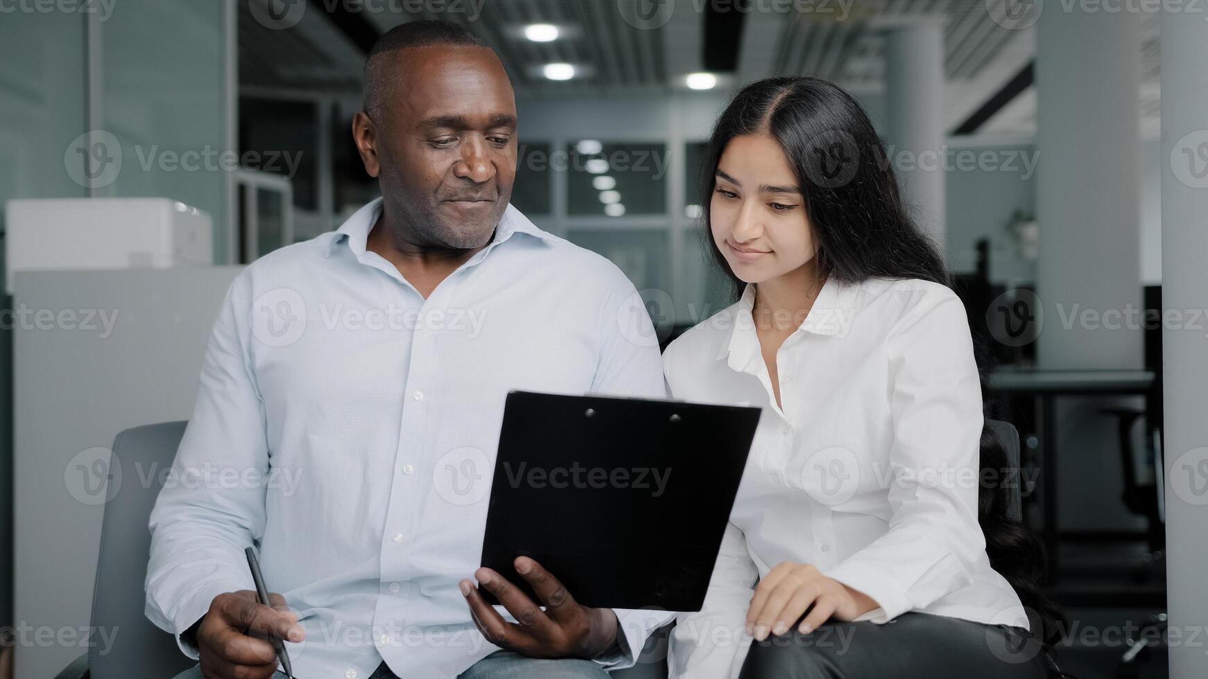 Diverse business partners sit in office discuss paper instruction african businessman show documentation in folder tablet to arabian female colleague explain insurance contract benefits negotiating photo