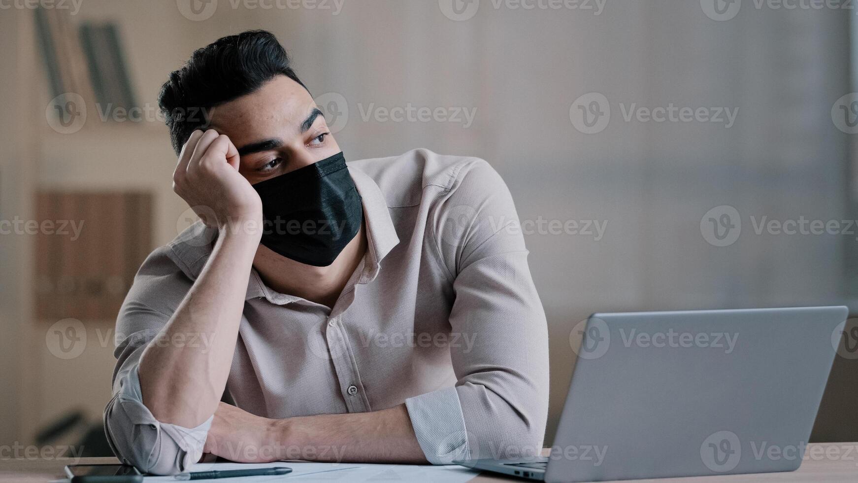 Bored sad hispanic worker young business man in medical mask tired unmotivated employee disinterested in dull distant work on computer lazy male arab guy feeling lack of motivation boredom in office photo