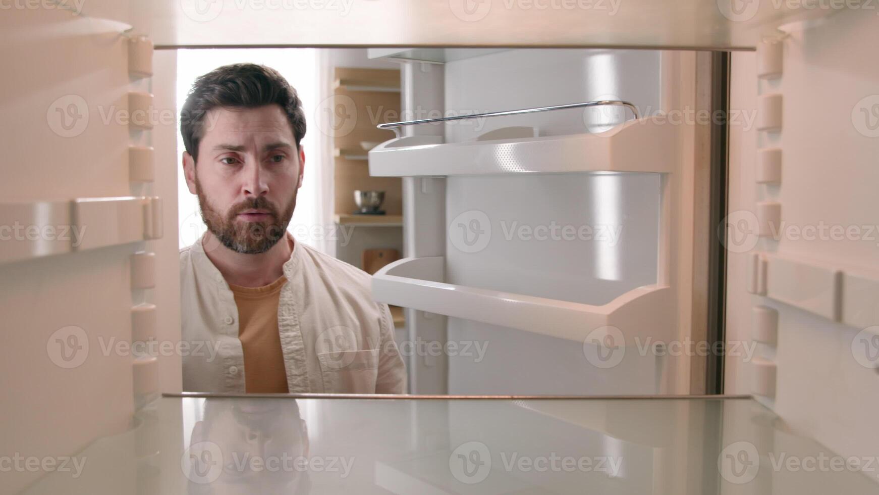 Hungry Caucasian bachelor man looking for food in empty fridge finding no meal products feeling sad disappointed poor unemployment need delivery service POV point of view from inside refrigerator photo