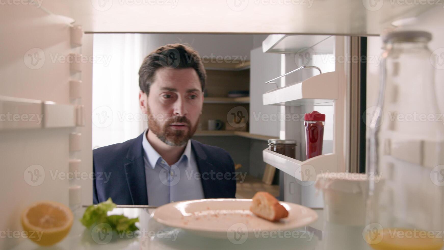 Hungry adult Caucasian businessman entrepreneur man after work open empty refrigerator with half lemon, lettuce leaf, bottle of milk and slice of bread eat crumbs POV point of view from inside fridge photo
