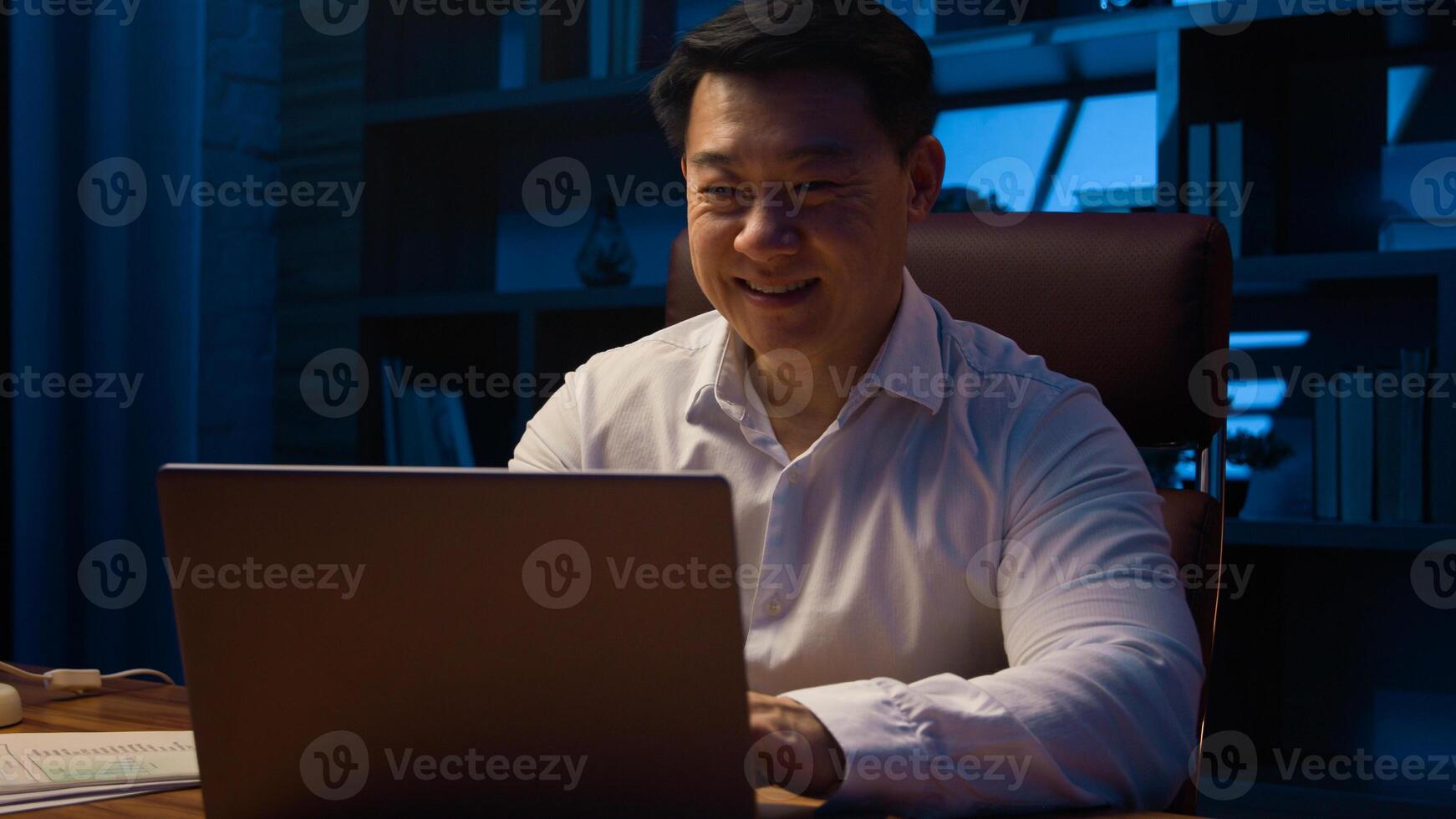Middle-aged happy smiling businessman working with laptop and documents at dark night office Asian Korean man financial analyst accounting business using computer app at nighttime insomnia deadline photo