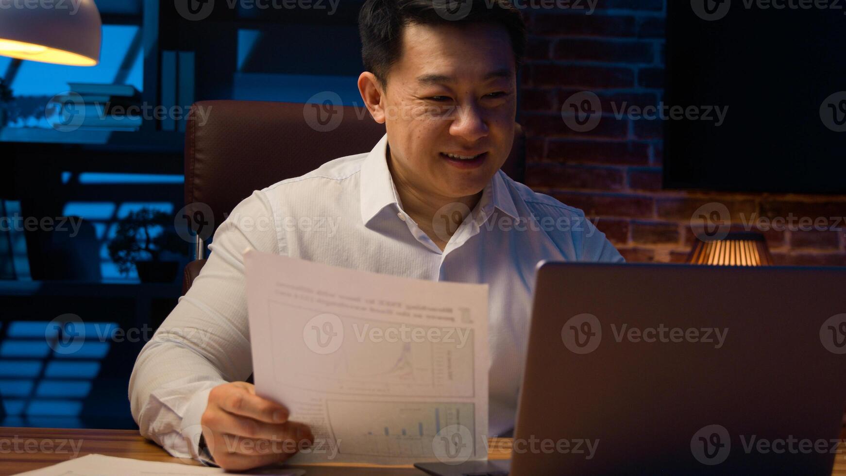 Middle-aged happy smiling businessman working with laptop and documents at dark night office Asian Korean man financial analyst accounting business using computer app at nighttime insomnia deadline photo