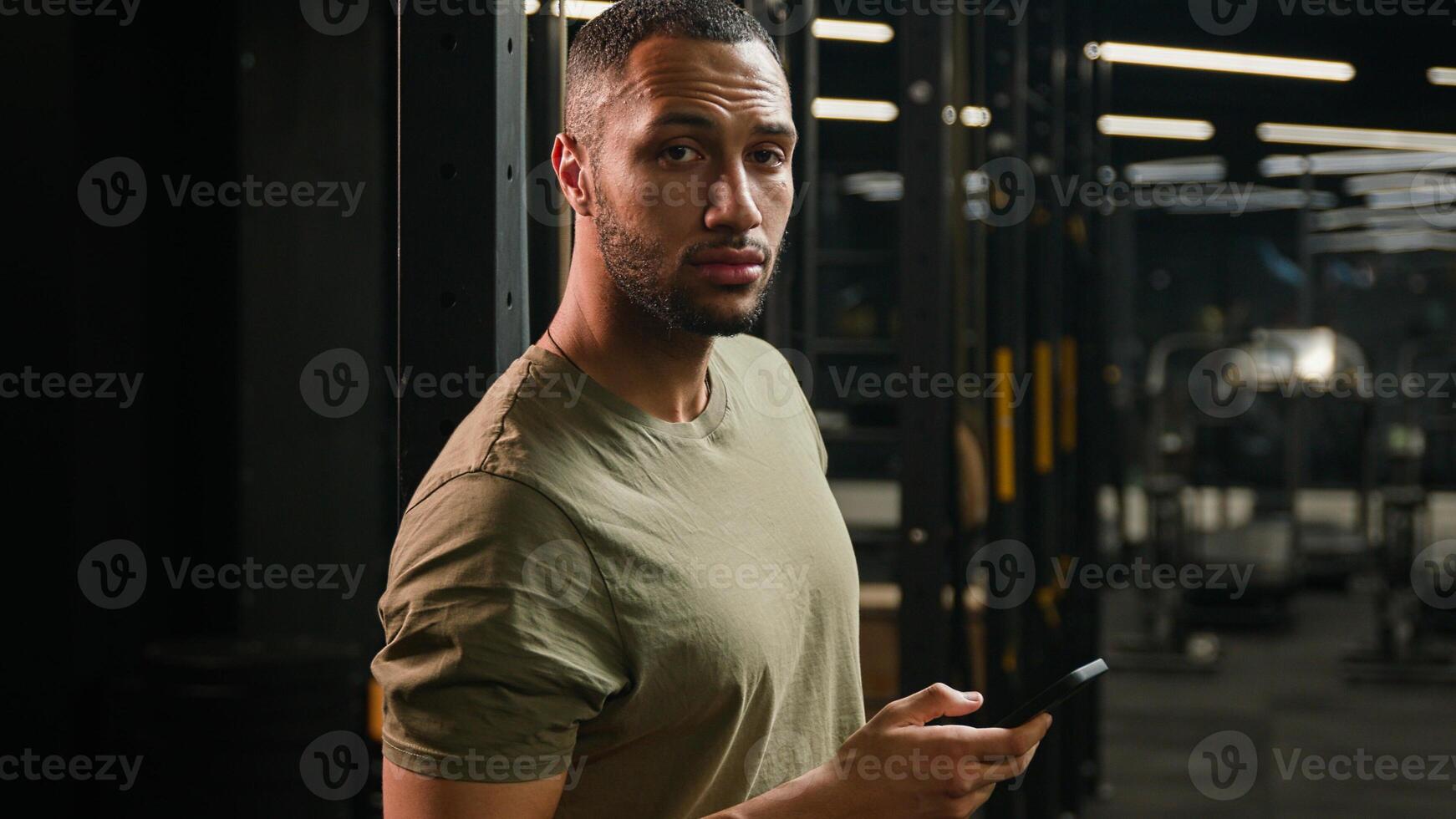 African American sportsman using mobile phone male fitness trainer man athlete relax in gym after workout exercise sport break browsing smartphone technology internet messaging calm looking at camera photo