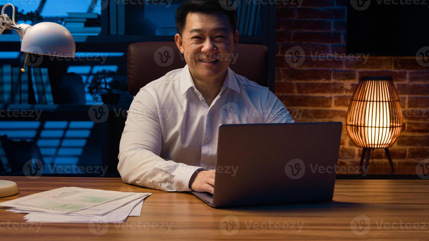 Middle-aged happy smiling businessman working with laptop and documents at dark night office Asian Korean man financial analyst accounting business using computer app at nighttime insomnia deadline photo