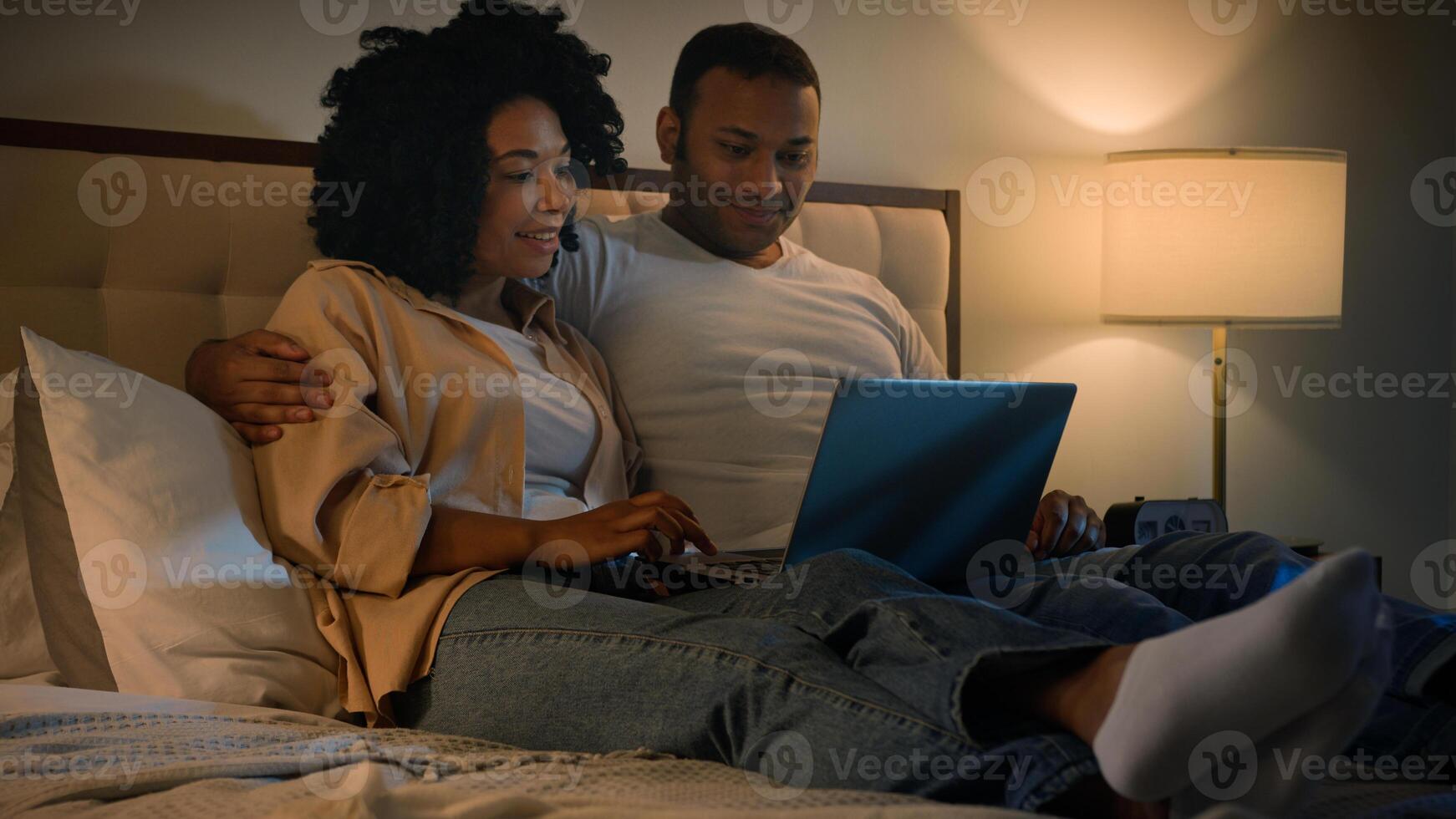 Happy family African American married couple husband man and wife woman on bed at night evening browsing net laptop using computer talking shopping app in internet choose film movie in online service photo