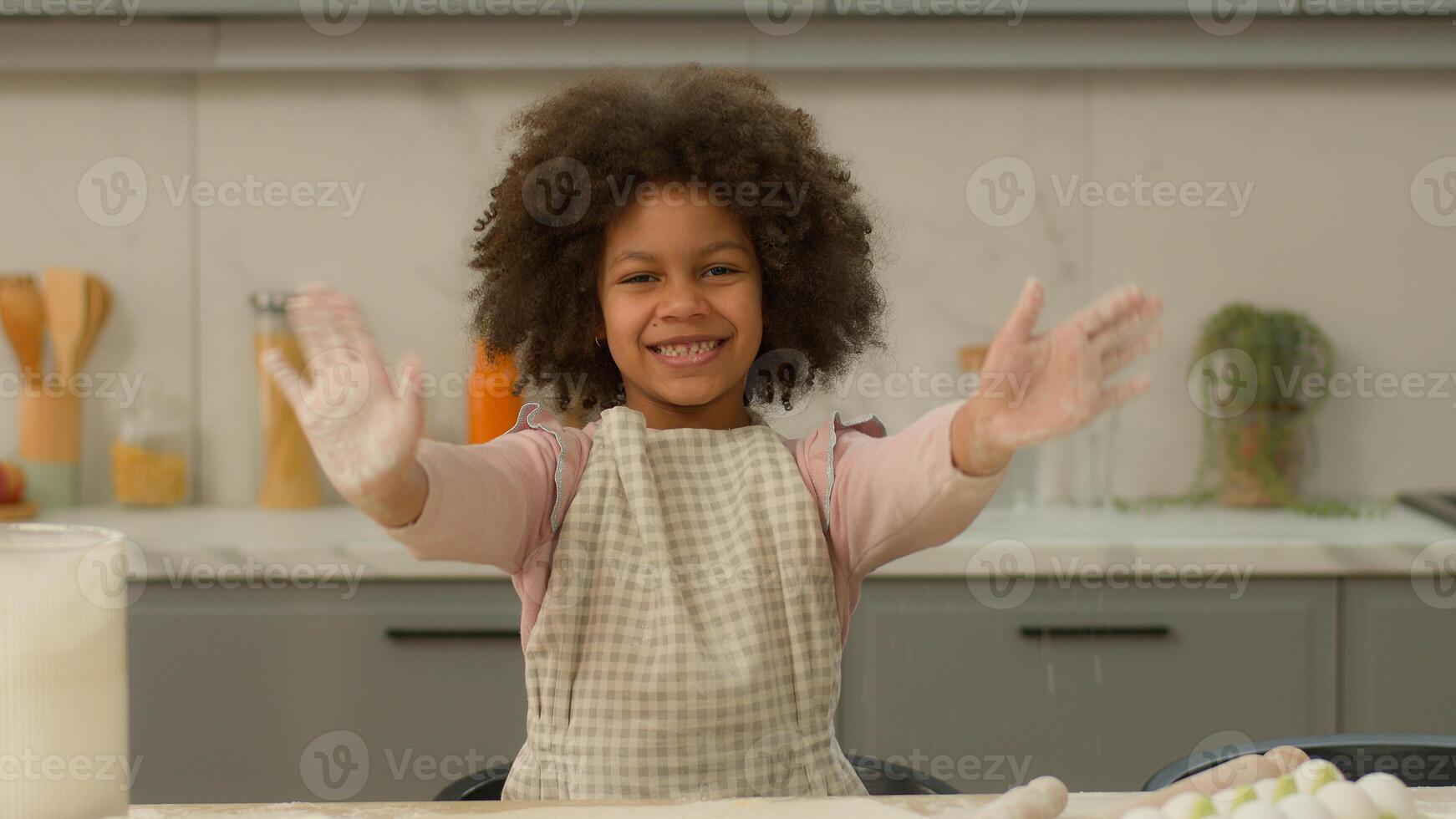 African American happy smiling positive cheerful ethnic little girl family adopted child kid daughter alone in apron waving hello smile greeting at camera in home kitchen learning cooking cook baking photo