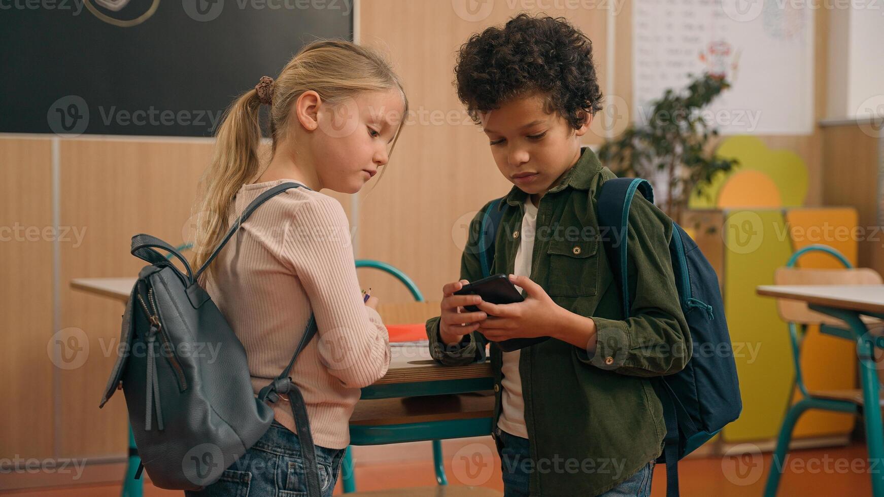 African and Caucasian little elementary school children multiracial diverse girl and boy kids pupils schoolchildren class study in classroom friendly talking using mobile phone holding smartphone talk photo
