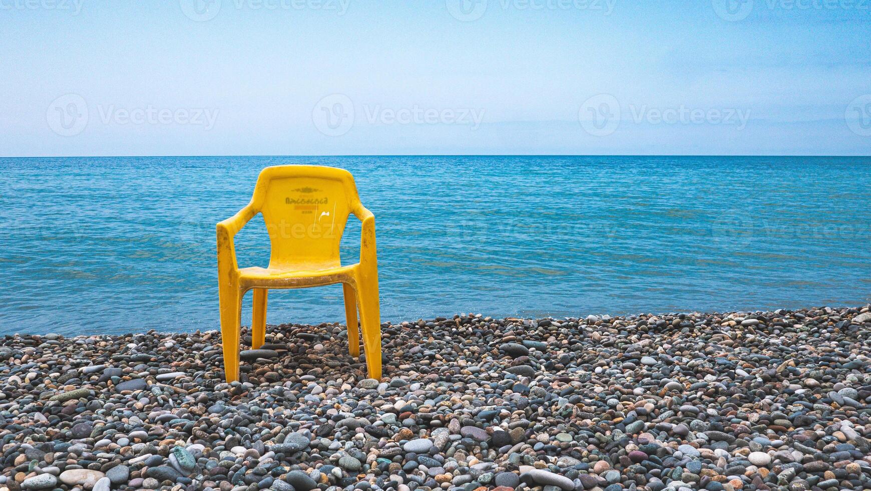 A yellow plastic chair on a stone beach in front of calm sea minimalist contrast photo