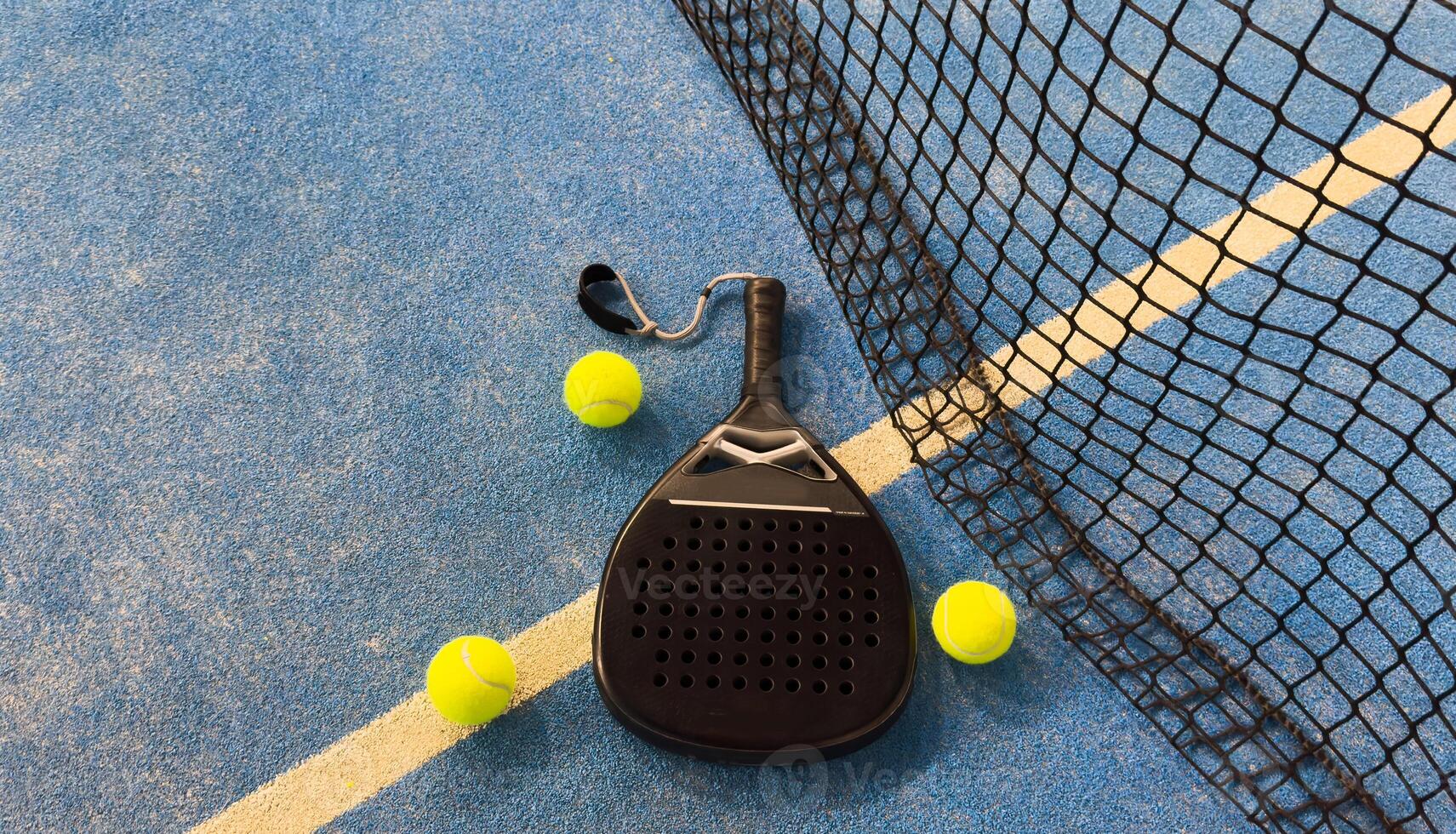 Padel racket and yellow ball behind net on a green court grass turf outdoors at sunset. Paddle is a racquet game. Professional sport concept with space for text photo